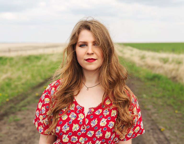 image of a woman in a red dress standing on an outside gravel road looking directly at the camera