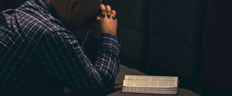 A man sits with his hands clasped in prayer beside an open Bible.