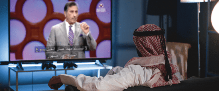 A Middle Eastern man in traditional attire watches a televised sermon by Dr. Michael A. Youssef from his living room.
